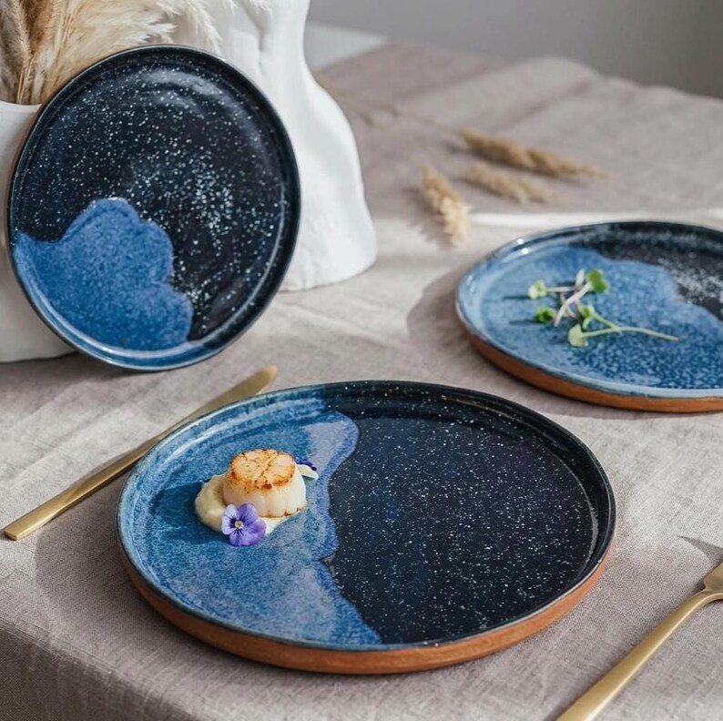 May include: A set of three ceramic plates with a speckled navy blue and light blue design. The plates have a unique wave-like pattern and a brown rim. One plate is garnished with food and a gold knife.