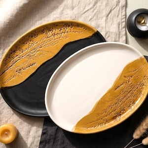 May include: Two oval ceramic platters featuring a two-tone design. One platter has a matte black base with a mustard yellow section embossed with a botanical grass pattern. The second platter has a white base with a matching mustard yellow section.
