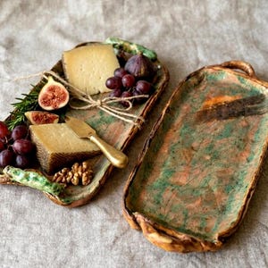 Pode incluir: Duas travessas retangulares de cerâmica, uma cheia de queijo, uvas, figos e nozes. A outra travessa está vazia. As travessas têm um design rústico com tons verdes e castanhos. Uma faca de queijo dourada repousa na travessa cheia.