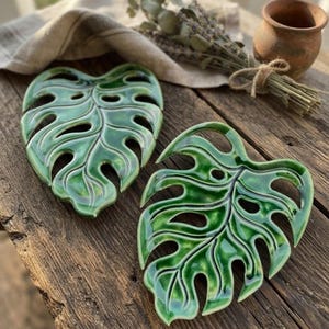 Puede incluir: Dos salvamanteles de cerámica de color verde oscuro brillante con forma de hojas de monstera. Cada pieza presenta recortes intrincados y venas en relieve, imitando la estructura natural de una hoja de planta tropical con un acabado liso.