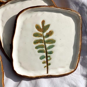 May include: Square ceramic plate with a white surface and a brown rim. The plate features a green and brown leaf design in the centre. The plates are stacked on top of each other.