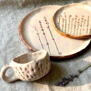May include: A ceramic mug and two plates with irregular, hand-formed edges. The off-white surfaces feature hand-painted lavender sprigs in shades of purple and green. A single dried lavender stem lies nearby on the textured fabric surface.