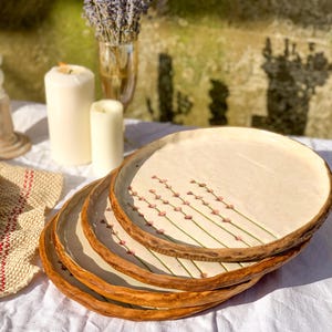 May include: A stack of four handmade ceramic plates. Each plate has a cream-colored center with a floral design and a brown, textured rim. The plates are stacked on a white tablecloth.