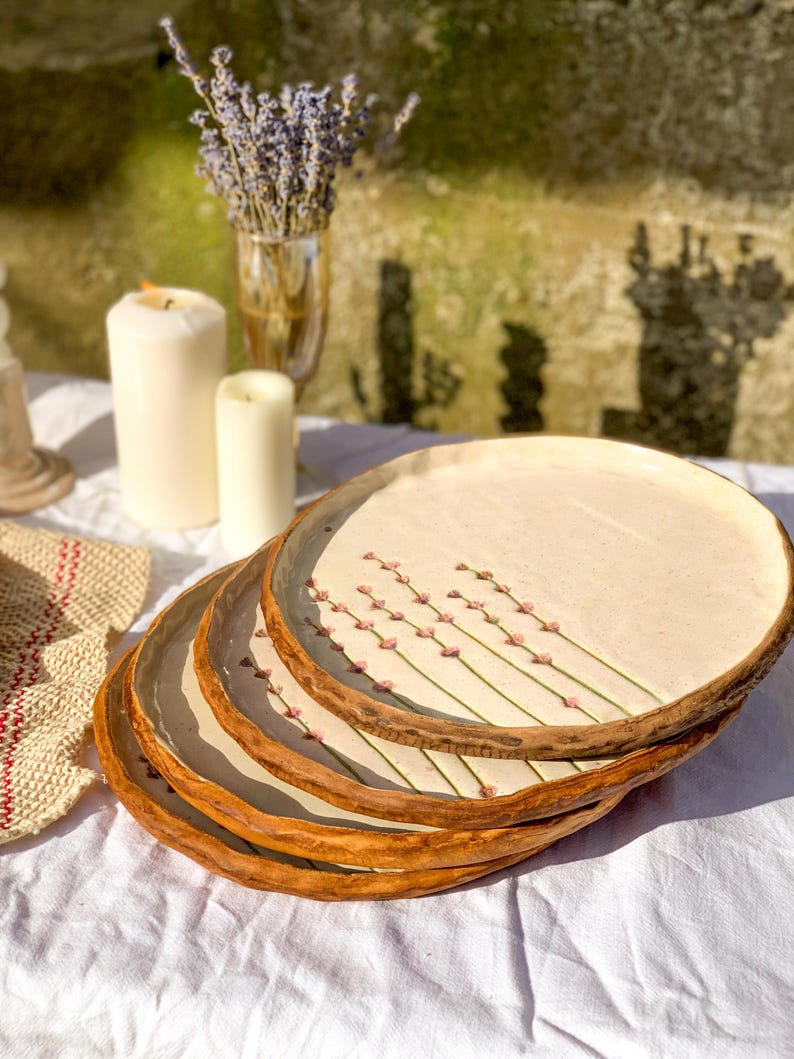May include: A stack of four handmade ceramic plates. Each plate has a cream-colored center with a floral design and a brown, textured rim. The plates are stacked on a white tablecloth.