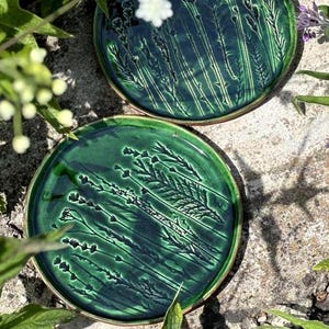 Può includere: Due piatti circolari in ceramica con una smaltatura verde scuro lucida. Ogni piatto presenta un dettagliato motivo botanico in rilievo di steli di grano e fiori selvatici impressi sulla superficie, creando un aspetto strutturato.