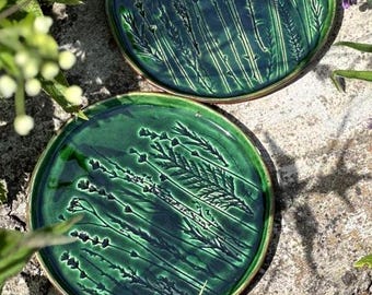 May include: Two circular ceramic plates featuring a glossy, deep green glaze. Each plate displays a detailed, embossed botanical pattern of wheat stalks and wildflowers pressed into the surface, creating a textured, relief-like appearance.