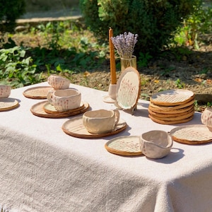 May include: A collection of handmade ceramic tableware, including cups, saucers, and plates, arranged on a linen tablecloth. The pieces are in a natural, off-white color with textured surfaces and a rustic aesthetic. A candle and a vase of lavender are also present.
