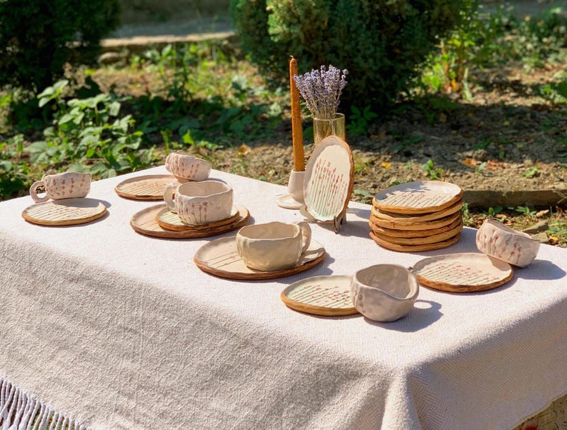 May include: A collection of handmade ceramic tableware, including cups, saucers, and plates, arranged on a linen tablecloth. The pieces are in a natural, off-white color with textured surfaces and a rustic aesthetic. A candle and a vase of lavender are also present.