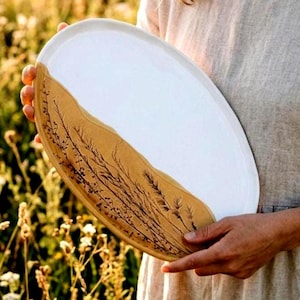 May include: An oval ceramic platter featuring a two-tone design. One half is finished in a smooth, glossy white glaze, while the other half displays a natural, unglazed tan clay surface decorated with detailed black botanical illustrations.