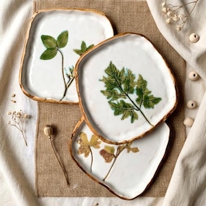 May include: Three square ceramic plates with white surfaces and gold rims. Each plate features a pressed botanical design, including a four-leaf clover and fern. The plates are arranged on a neutral linen surface.
