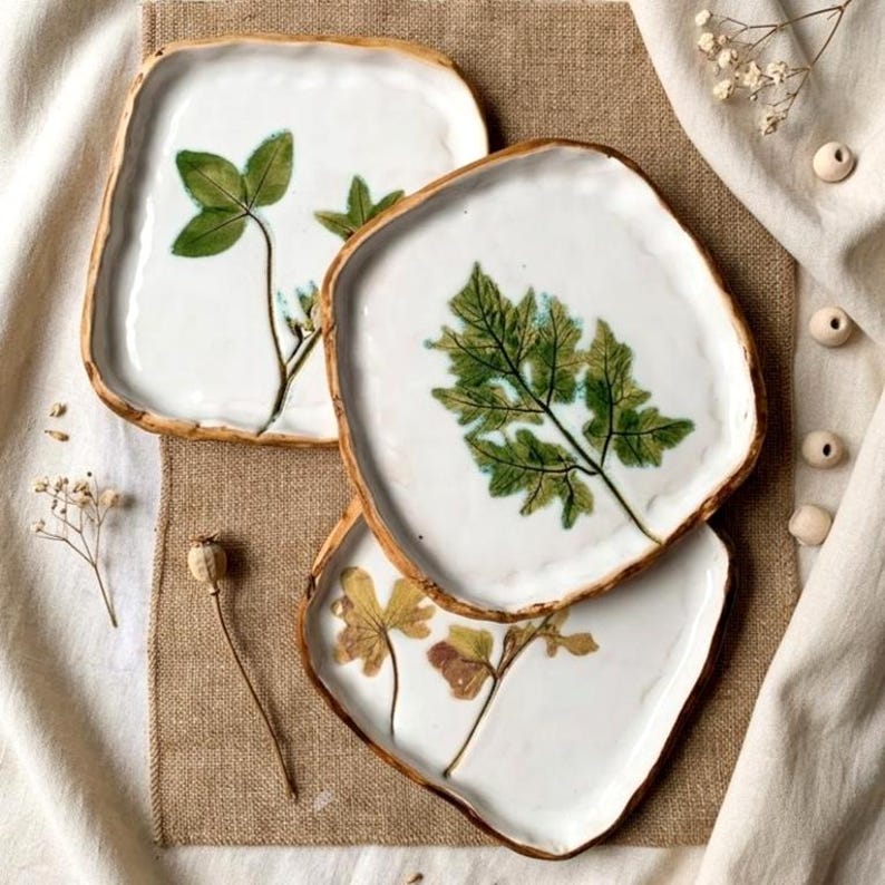 May include: Three square ceramic plates with white surfaces and gold rims. Each plate features a pressed botanical design, including a four-leaf clover and fern. The plates are arranged on a neutral linen surface.