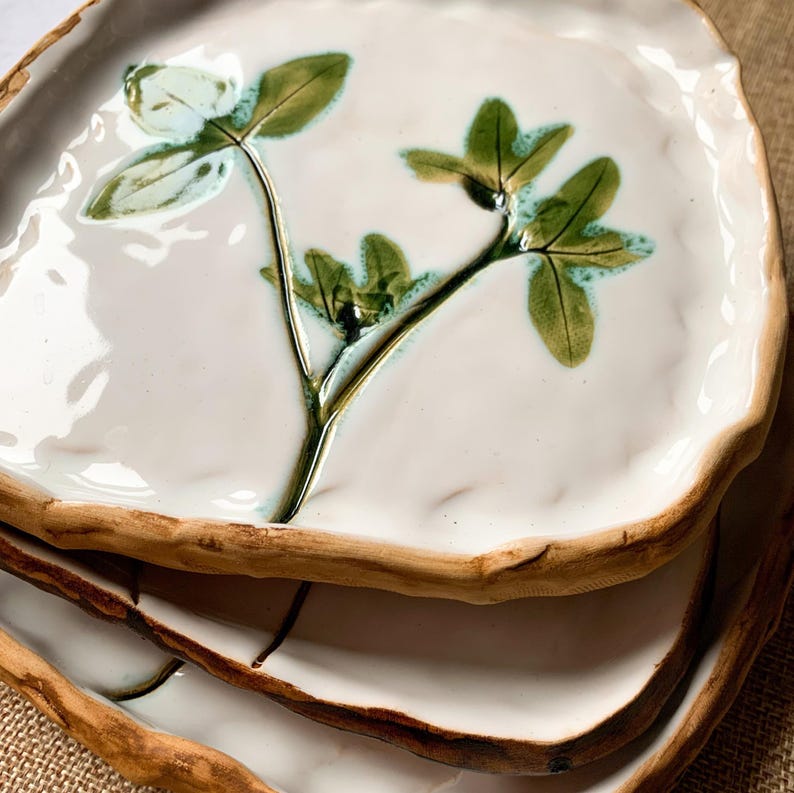 May include: A stack of handmade ceramic plates. Each plate has a white glazed surface with a green botanical design. The rims are a natural, unglazed brown colour, and the plates have an organic, irregular shape.