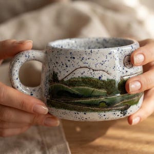 May include: A ceramic mug with a white base and blue speckles, featuring a hand-painted landscape design of green rolling hills and a mountain range. The mug has a wide, rounded handle and a smooth, glossy finish across the entire exterior surface.