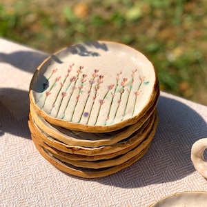 May include: A stack of handmade ceramic plates. Each plate features a cream-colored surface with a floral design of lavender-colored flowers and green stems. The rims are a warm brown color. The plates are stacked on a light-colored surface.