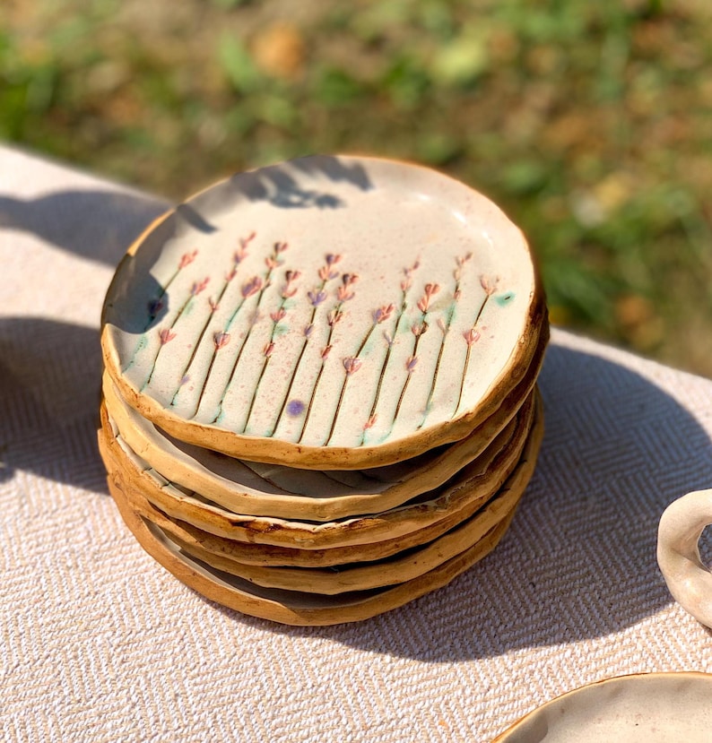 May include: A stack of handmade ceramic plates. Each plate features a cream-colored surface with a floral design of lavender-colored flowers and green stems. The rims are a warm brown color. The plates are stacked on a light-colored surface.