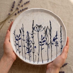 May include: A round, speckled ceramic plate with a hand-painted botanical design. The plate features tall, slender stems and leaves in shades of blue and black against a white background. The plate is held by two hands.