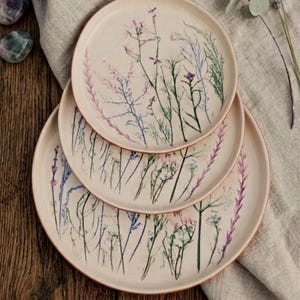 Pode incluir: Três pratos de cerâmica bege empilhados, cada um adornado com delicados desenhos florais em roxo, azul e verde. Os pratos têm uma borda ligeiramente elevada e uma aparência rústica e artesanal, perfeitos para servir comida.