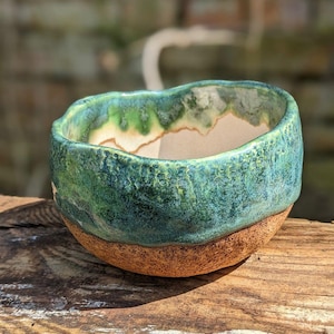 May include: A handcrafted ceramic bowl with a textured, turquoise and green glazed exterior. The bowl's base is a natural, unglazed brown. The rim has an organic, uneven shape. The bowl is likely used for serving or display.
