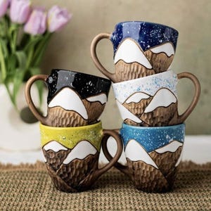 Puede incluir: Una pila de seis tazas de cerámica con diseños de montañas. Cada taza presenta una combinación de colores única, incluyendo azul, negro, blanco, amarillo y marrón. Las tazas tienen asas marrones y un aspecto texturizado y tallado.