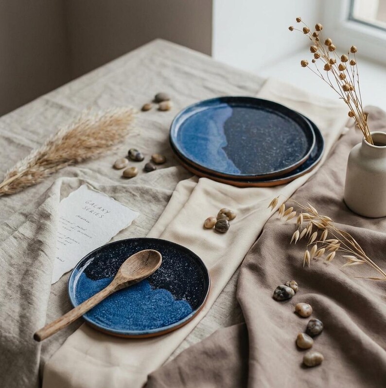 May include: A set of ceramic plates with a blue and black galaxy design. One plate has a wooden spoon. The plates are on a beige tablecloth with small stones and a vase with dried flowers. The text "GALAXY SERIES" is on a piece of paper.