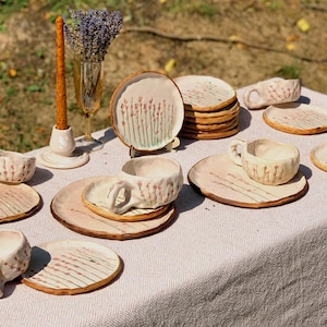May include: A collection of handmade ceramic tableware, including cups, saucers, and plates, arranged on a table. The pieces feature a light beige color with a floral design in pink and green. A tall, brown candle and a vase of lavender are also present.