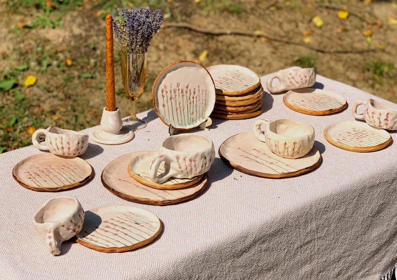 May include: A collection of handmade ceramic tableware, including cups, saucers, and plates, arranged on a table. The pieces feature a light beige color with a floral design in pink and green. A tall, brown candle and a vase of lavender are also present.