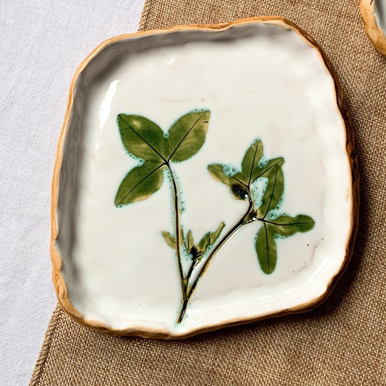May include: Square ceramic plate with a white surface and a four-leaf clover design. The clover is green with blue-green accents. The plate has a textured, light brown rim. The plate is on a brown surface.