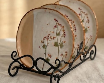 May include: Three round ceramic plates with uneven edges, featuring pressed dried flowers in shades of pink and green on a white glazed surface. The plates are held upright in a black metal stand with decorative scrollwork and a leaf detail.