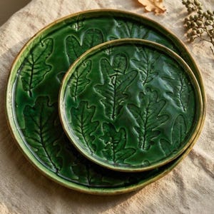 May include: Two green ceramic plates with a raised oak leaf design. The plates are different sizes and have a glossy finish with a gold-colored rim. The leaves are detailed and create a textured surface.