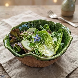 Puede incluir: Un cuenco de cerámica con exterior marrón texturizado y borde verde ondulado brillante, lleno de una ensalada fresca de hojas de lechuga verde, rodajas finas de rábano, brotes y una pequeña flor de borraja azul en el centro del cuenco.