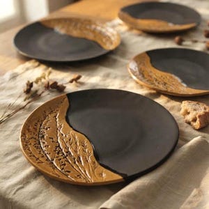 May include: A set of round ceramic plates featuring a matte black finish on one side and a textured, golden-brown botanical relief pattern on the other. The plates have a smooth, dark center and a raised, detailed leaf design along the curved edge.