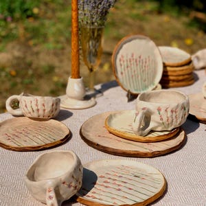 May include: A collection of handmade ceramic tableware, including cups, saucers, and plates, arranged on a table. The pieces feature a light beige base with a floral design in pink and green. A tall orange candle and a vase of lavender are also present.