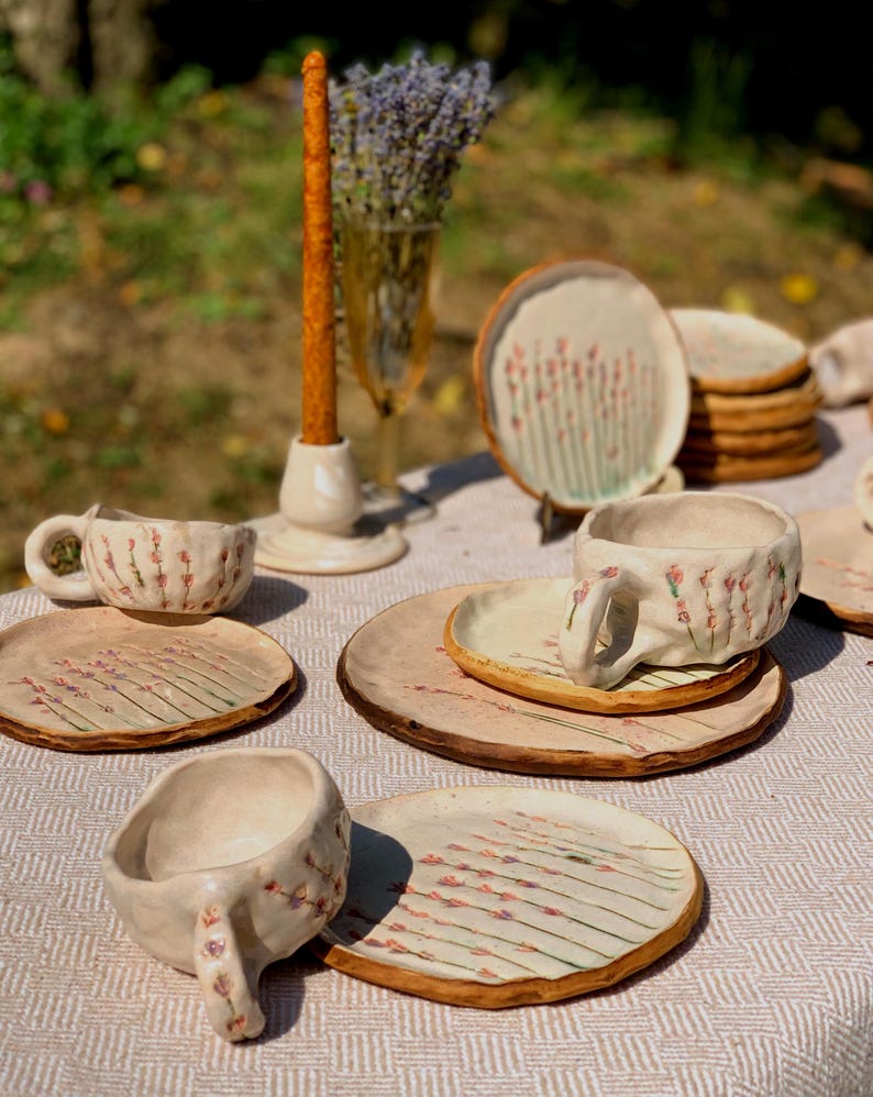 May include: A collection of handmade ceramic tableware, including cups, saucers, and plates, arranged on a table. The pieces feature a light beige base with a floral design in pink and green. A tall orange candle and a vase of lavender are also present.