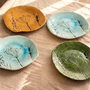 May include: Four round ceramic plates featuring embossed botanical designs of wildflowers and wheat stalks. The set includes one mustard yellow plate, two light blue plates, and one sage green plate, all with a textured, rustic finish.