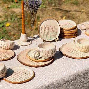 May include: A collection of handmade ceramic tableware, including plates and cups, arranged on a table. The plates and cups are beige with a floral design in pink and green. A candle and a vase with lavender are also on the table.