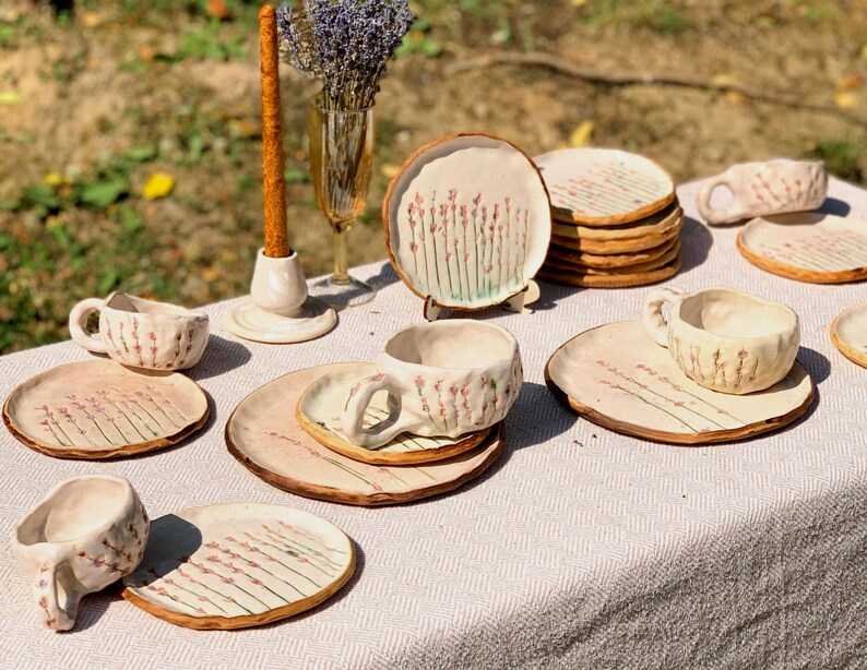 May include: A collection of handmade ceramic tableware, including plates and cups, arranged on a table. The plates and cups are beige with a floral design in pink and green. A candle and a vase with lavender are also on the table.