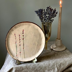 May include: A ceramic plate with a lavender design, displayed on a small stand. The plate has a light beige center with a brown rim. A lit candle and a vase of dried lavender are also in the image.