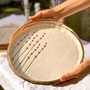 May include: A stack of handmade ceramic plates. Each plate has a cream-colored surface with a brown rim. Delicate lavender sprigs are painted on the surface. The plates are held in front of a white tablecloth.