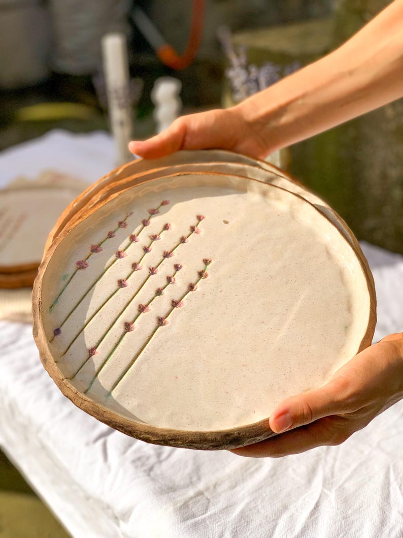 May include: A stack of handmade ceramic plates. Each plate has a cream-colored surface with a brown rim. Delicate lavender sprigs are painted on the surface. The plates are held in front of a white tablecloth.