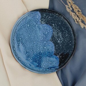 May include: A round ceramic plate with a two-tone design. The plate features a textured, speckled blue surface that transitions to a dark blue and black area with white speckles. The plate is set against a neutral background.