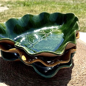 May include: A stack of three ceramic bowls shaped like large green leaves. Each bowl features a glossy dark green glaze with visible embossed vein patterns on the interior surface and a wavy, scalloped edge. The undersides are a natural tan color.