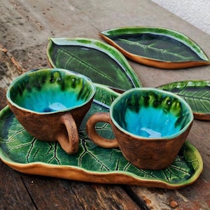 Pode incluir: Conjunto de chávenas e pires de cerâmica feitos à mão. As chávenas têm exterior castanho e interior turquesa. Os pires têm formato de folha com esmalte verde. O conjunto inclui duas chávenas e dois pires.