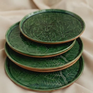 Puede incluir: Una pila de cuatro platos redondos de cerámica con un esmalte verde oscuro brillante. Cada plato presenta un patrón botánico en relieve distinto, incluyendo hojas de roble y frondas de helecho, con la arcilla de terracota natural visible.