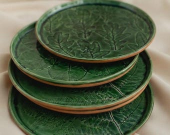 May include: A stack of four round ceramic plates with a glossy dark green glaze. Each plate features a distinct embossed botanical pattern, including oak leaves and fern fronds, with the natural terracotta clay visible along the outer edges.