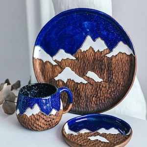 May include: A ceramic tableware set featuring a large plate, a mug, and a small dish. The pieces have a blue and white mountain design over a brown textured base. The mug has a handle and a speckled interior.