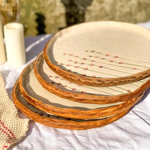 May include: A stack of four handmade ceramic plates. The plates have a cream-colored center with a floral design of pink buds on green stems. The rims are a textured, warm brown. The plates are stacked on a white surface.