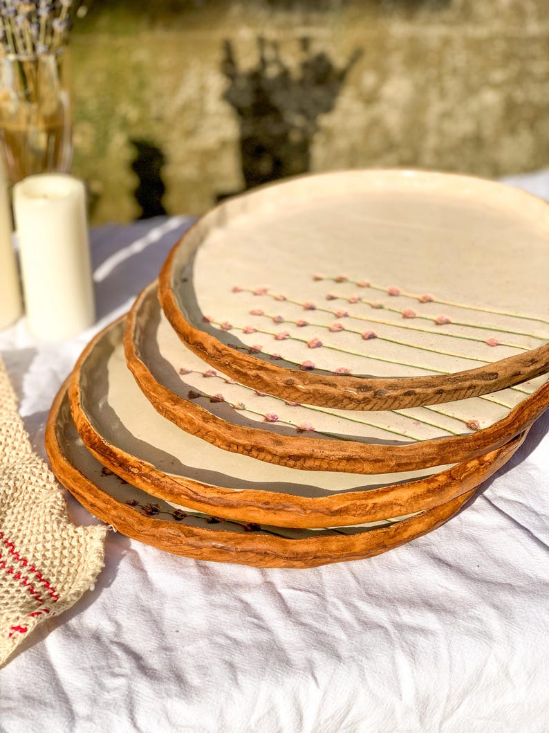May include: A stack of four handmade ceramic plates. The plates have a cream-colored center with a floral design of pink buds on green stems. The rims are a textured, warm brown. The plates are stacked on a white surface.