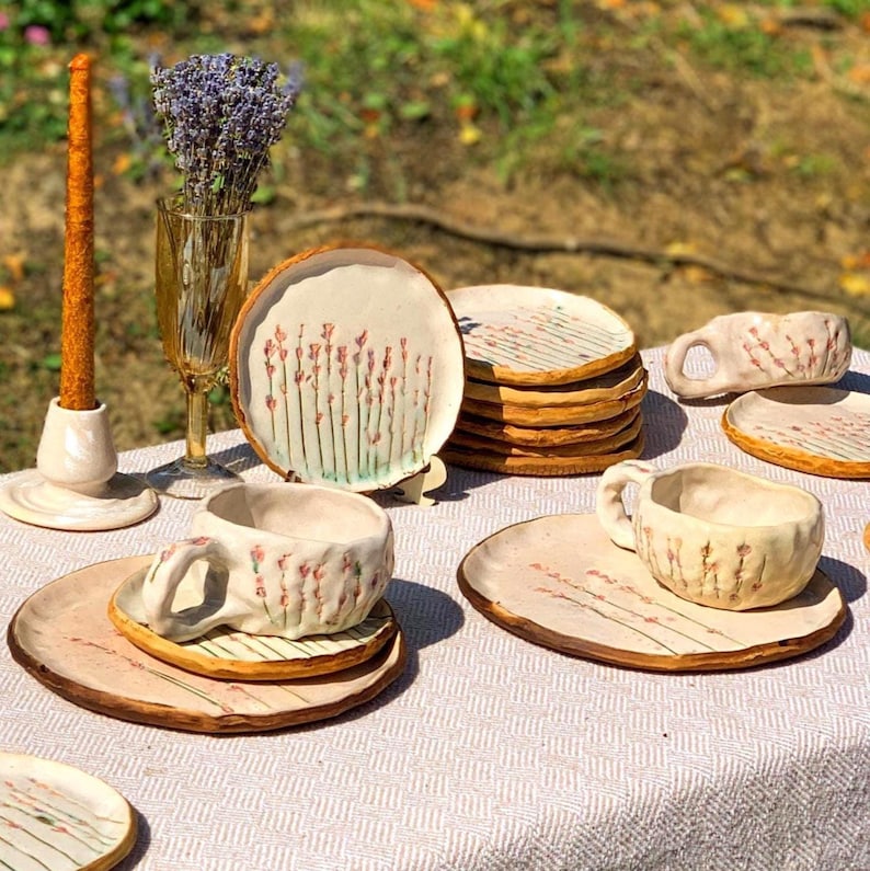 May include: A collection of handmade ceramic tableware, including plates, cups, and saucers, arranged on a table. The pieces feature a cream-colored base with a brown rim and delicate floral designs in pink and green. A candle and a vase of lavender are also present.