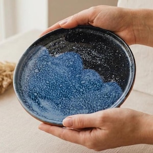 May include: A handmade ceramic plate with a unique design. The plate features a dark blue and black glaze with a textured, speckled finish. The lower half of the plate is a lighter blue. The plate is held by two hands.