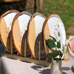 Op de afbeelding: Vier decoratieve keramische borden met een natuurlijke houten rand, tentoongesteld in een houten rek. Elk bord heeft een wit en bruin kleurenschema met bloemaccenten. Een kleine vaas met groen en een roze roos staat op de voorgrond.
